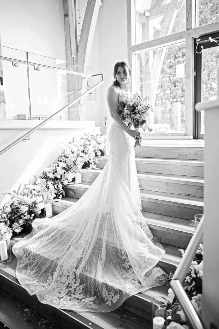 Portrait black and white photo of the bride standing on a set of stairs with her wedding dress trailing with flowers eiather side of the stairs.
