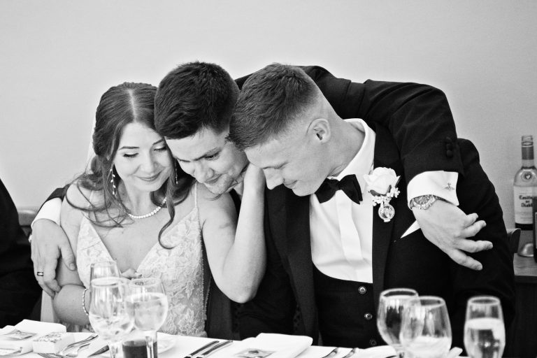 B&W storytelling photo of the best man hugging the bride and groom.